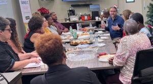 Photo of a group of people having a discussion around a table, sharing food and taking notes.