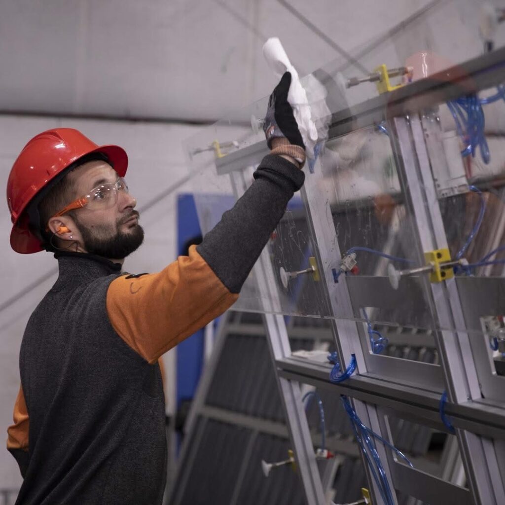 A worker in a hard hat and safety glasses wipes down a glass pane.