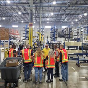 Workers in high-visibility vests standing in a warehouse face away from the viewer.