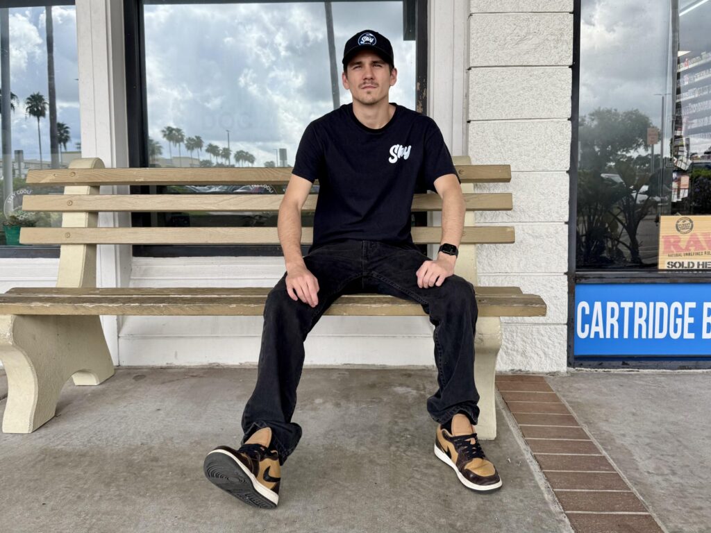 A person in a Sky branded cap and t-shirt sits on a bench outside a vape shop.