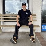 A person in a Sky branded cap and t-shirt sits on a bench outside a vape shop.