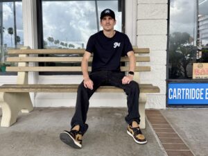 A person in a Sky branded cap and t-shirt sits on a bench outside a vape shop.