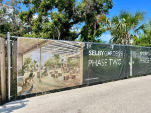 A banner on a chain-link fence separating a road from a natural area advertises "Selby Gardens Phase Two: with a lifelike rendering of a large porch.