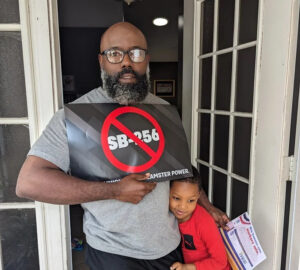 Standing on the sidewalk with an umbrella, a person holds a sign with a red circle and line through text that reads "SB-256" while a child hugs them.