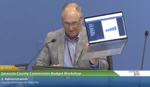 Tom Knight speaking while holding up a laptop which displays a bar graph.