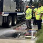 Workers wearing hi-vis overshirts sandblast artwork off a sidewalk.