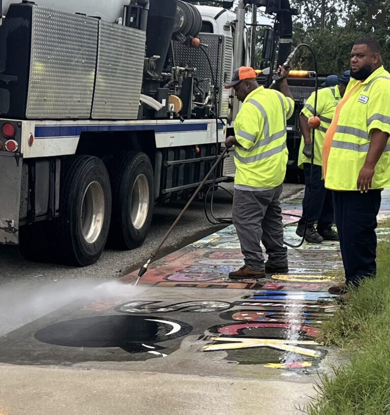 Workers wearing hi-vis overshirts sandblast artwork off a sidewalk.
