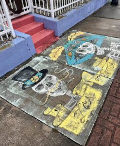Sidewalk art of two well-dressed skeletons with a banner that reads "Los Muertos".