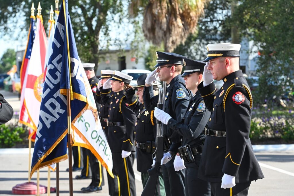Firefighters and police officers in ceremonial uniforms salute in front of U.S, Florida, Sherriff's Office and police department flags.