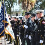 Firefighters and police officers in ceremonial uniforms salute in front of U.S, Florida, Sherriff's Office and police department flags.