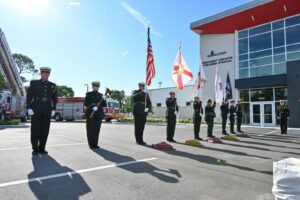 Firefighters and police officers in ceremonial uniforms bear U.S, Florida, Sherriff's Office and police department flags in front of a fire station.