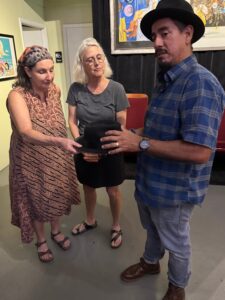 Oliver Dominguez, wearing a hat, shows off another hat to two interested people.
