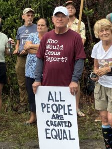 A demonstrator wearing a shirt that reads "Who Would Jesus Deport?" holds a handwritten sign that reads "all people are created equal".
