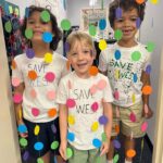 Photo of three elementary students standing in a hallway behind a beaded curtain and smiling. They wear shirts with "Save W.E.S." handwritten on them.
