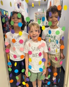 Photo of three elementary students standing in a hallway behind a beaded curtain and smiling. They wear shirts with "Save W.E.S." handwritten on them.