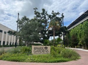 Sign that reads "Marie Selby Botanical Gardens Downtown Sarasota" surrounded by leafy and flowering plants.