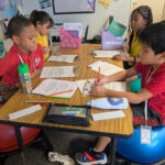 Photo of elementary students sitting around a table and chatting while working on worksheets.