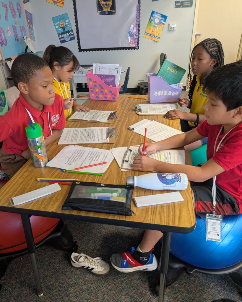 Photo of elementary students sitting around a table and chatting while working on worksheets.