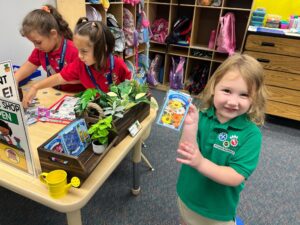 Photo of elementary students at a booth in a classroom selling plants and plant seeds for fake money. They wear polo shirts with an embroidered design that reads "Building the future" and "Wilkinson Elementary".