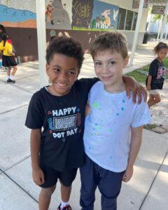 Photo of two elementary students standing outside a school and smiling. One has an arm around the other.