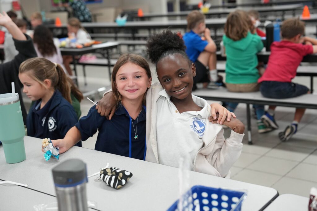 Photo of two elementary students sitting at a cafeteria table and smiling, each with an arm around the other's shoulder.