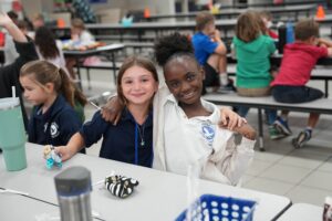 Photo of two elementary students sitting at a cafeteria table and smiling, each with an arm around the other's shoulder.