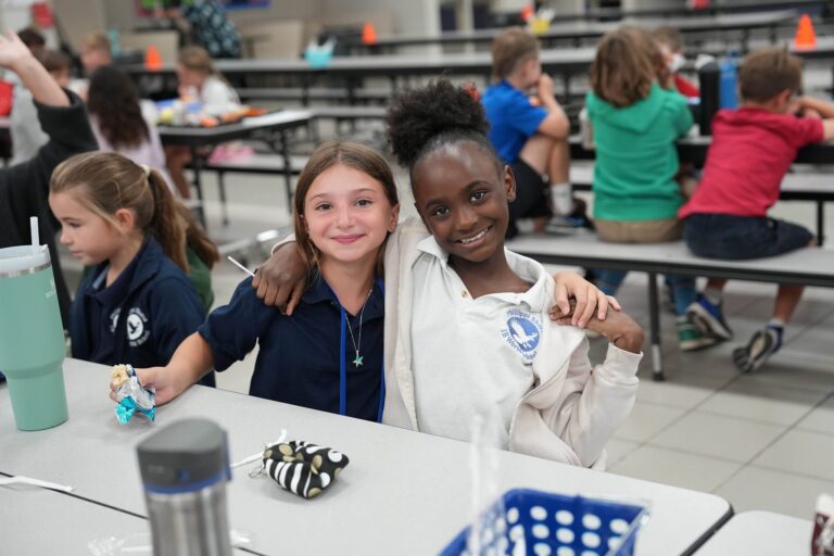 Photo of two elementary students sitting at a cafeteria table and smiling, each with an arm around the other's shoulder.
