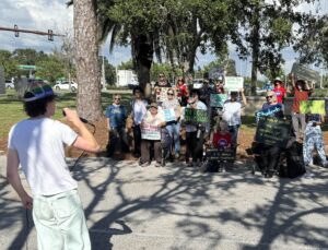 Photo of a speaker in a pride flag-colored cap addressing a crowd holding signs in defense of public schools.