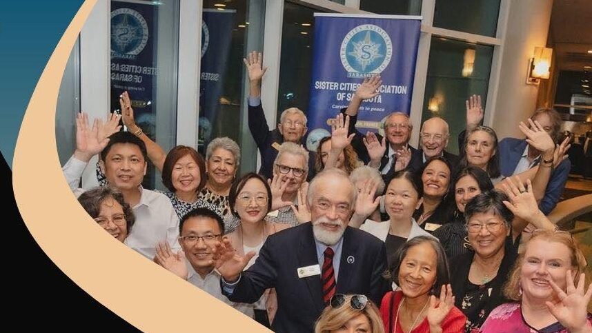 Photo of a crowd of people smiling in front of a Sister Cities Association of Sarasota banner.