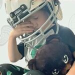 Photo of a child wearing a football helmet and clutching a stuffed bear with a pained expression.