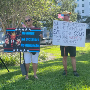 Two demonstrators holding signs. One has a crossed-out photo of Donald Trump wearing a crown and says "No Kings, No Dictators, No Oligarchs". The other one reads "'All that is necessary for the triumph of evil is that good men (and women) do nothing.' -Edmund Burke".