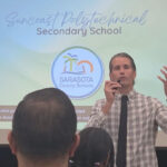 Terry Connor addressing a crowd in front of a projected presentation slide titled Suncoast Polytechnical Secondary School.