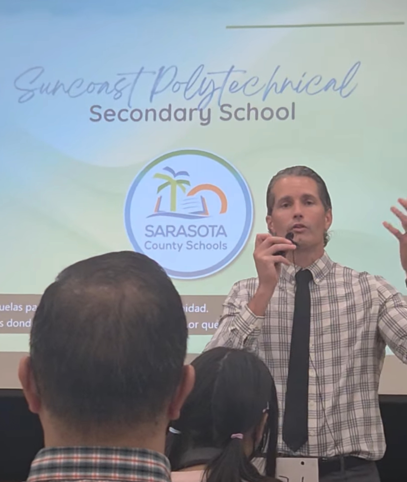 Terry Connor addressing a crowd in front of a projected presentation slide titled Suncoast Polytechnical Secondary School.