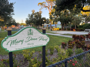 Photo of a sign reading "Mary Dean Park" with a playground and basketball court in the background.