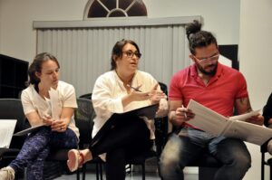 Three cast members sit in a row. Ivonne Batanero and Edward Ossa look at scripts. Perla Avalos gesticulates while speaking.