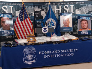A Homeland Security Investigations table with several firearms on display and three arrest photos captioned with the individuals' nationalities and alleged crimes.