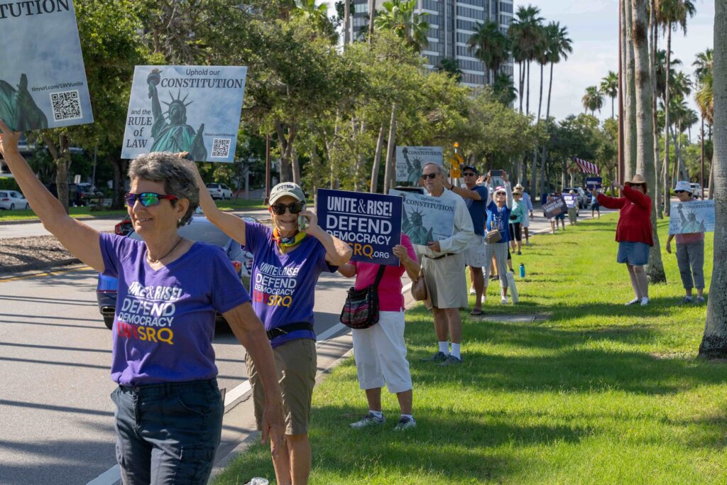Demonstrators near downtown Sarasota wear shirts and hold signs that read "Unite & rise! Defend democracy LWV SRW" and "Uphold our Constitution and the rule of law".