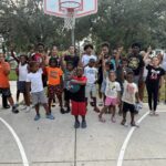 Children pose pointing upward in front of a basketball hoop in a park.