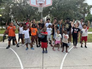Children pose pointing upward in front of a basketball hoop in a park.