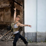 A person in a military vest with a Ukrainian flag emblem dancing near the wreckage of a building.