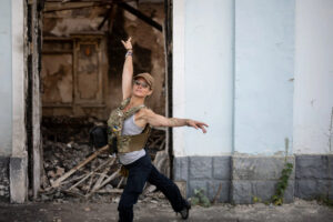 A person in a military vest with a Ukrainian flag emblem dancing near the wreckage of a building.
