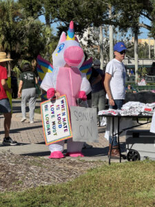 A demonstrator in an inflatable unicorn costume holds two signs that read "When hate is loud love must be louder" and "no kings".