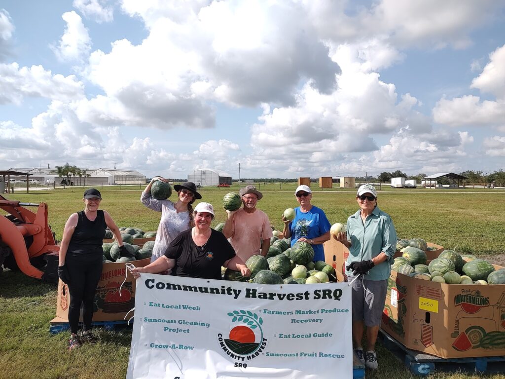 A group of people outdoors standing near crates of watermelons, holding a Community Harvest SRQ banner and smiling.