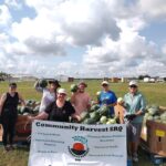 A group of people outdoors standing near crates of watermelons, holding a Community Harvest SRQ banner and smiling.