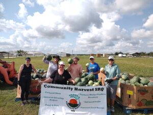 A group of people outdoors standing near crates of watermelons, holding a Community Harvest SRQ banner and smiling.