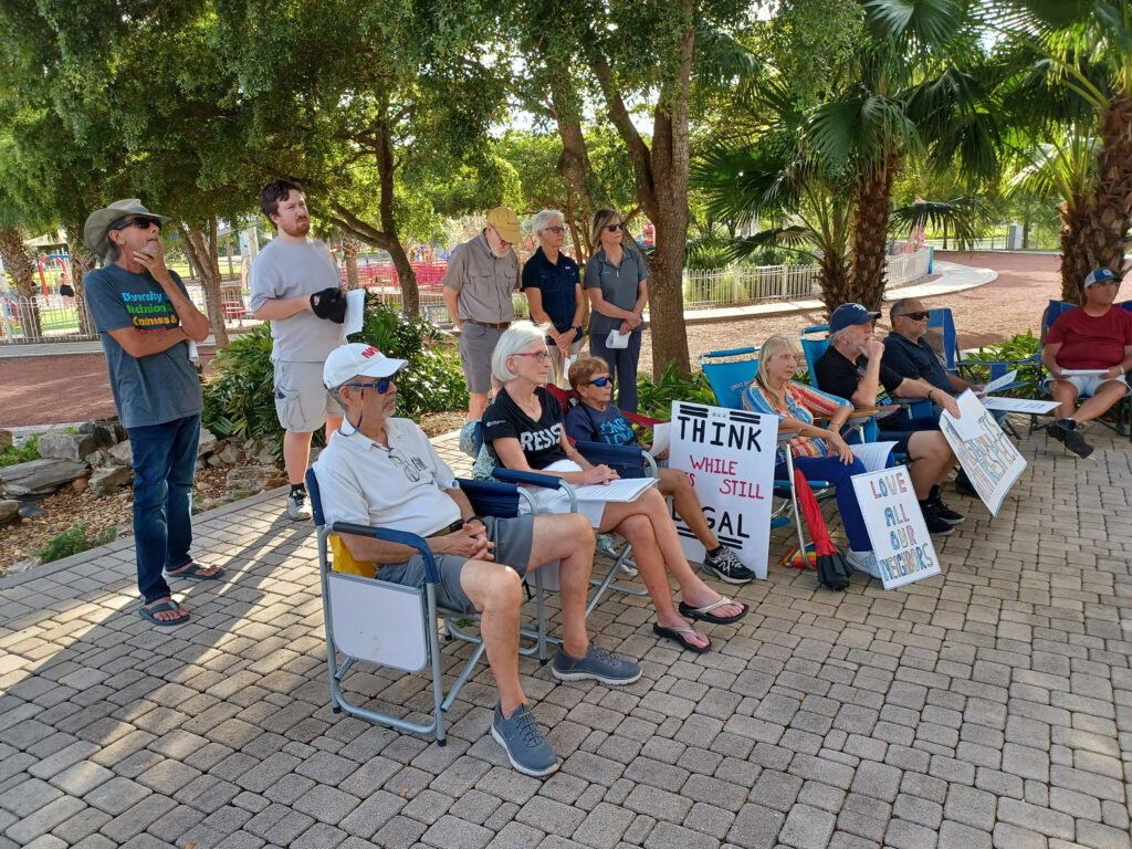 A group of demonstrators sit with handwritten signs with messages like "Think whyle it's still legal" and "Love all our neighbors".