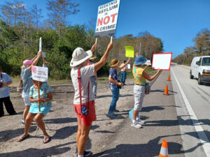 Demonstrators along the side of the road holding signs including "Seeking asylum is not a crime" and "Hands off the everglades".