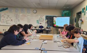 Middle school students work at a table in a classroom to make pipe cleaner models of DNA strands.