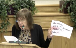 Robin Williams speaking at a podium holding a piece of paper that reads "Repeal co-location support SB424!"