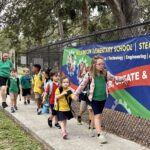 Elementary school students walk along a sidewalk near a Wilkinson Elementary School banner under adult supervision.